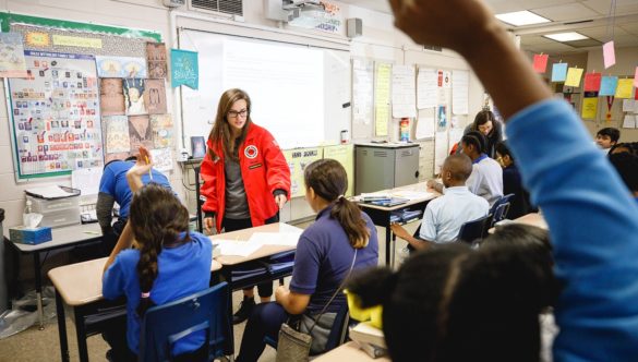 elementary students are in classroom with hands raised while AmeriCorps member rotates through and talks to students