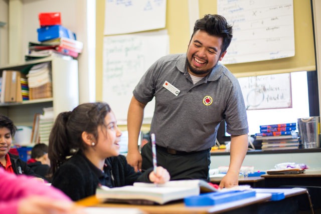 city year americorps member laughing with a student who is sitting at a desk and writing in her book