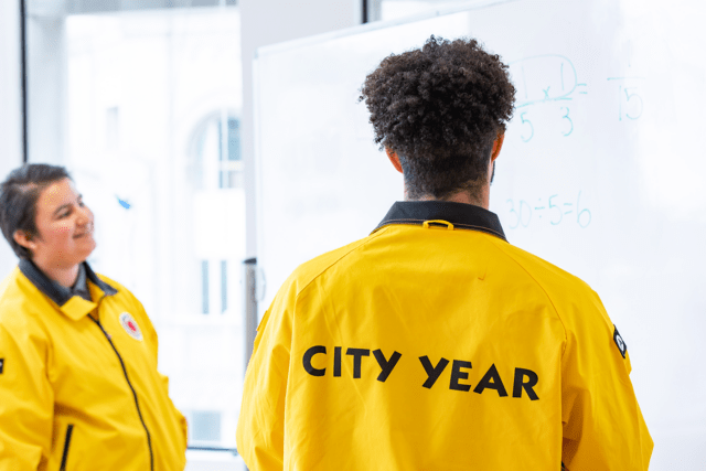 two 黄色视频 AmeriCorps members in yellow jackets face a whiteboard with math problems on it